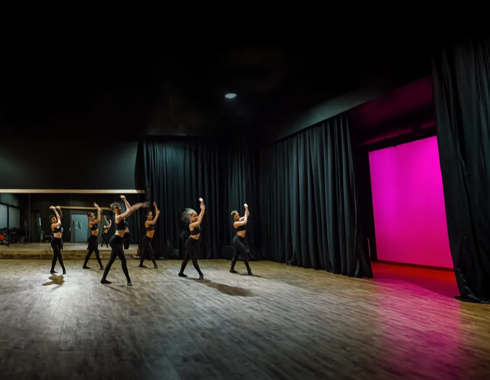 Six dancers in a line practising sharp vogue arm shapes on a wooden floor, a magenta backdrop behind them.