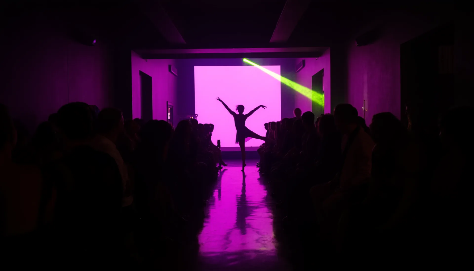 Looking down the runway in a dark club: one person stands at the far end in magenta backlight, the audience seated tight along both sides.