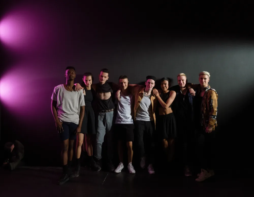 Eight dancers standing shoulder to shoulder in a dark rehearsal room, magenta light from the left.
